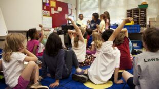 Skolbarn sittande på golvet i ett klassrum. Vuxen i bakgrunden. Flera barn räcker upp handen.