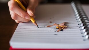 En hand som skriver med blyertspenna på randigt block, rester från vässad penna på pappret.