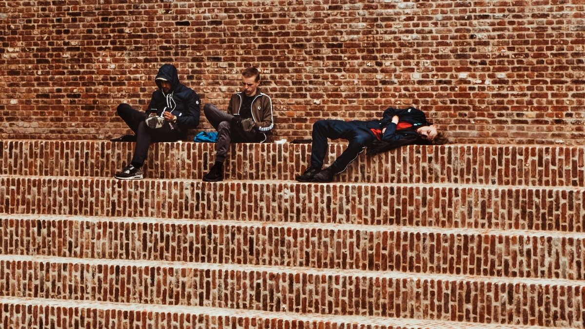 Tre ungdomar i svart i en tegelbeklädd trappa utomhus. En av personerna ligger och verkar sova.