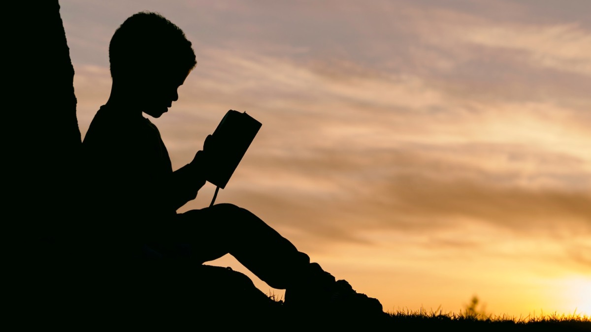 Barn, i mörk profil i solnedgång sittande mot träd, läser en bok.