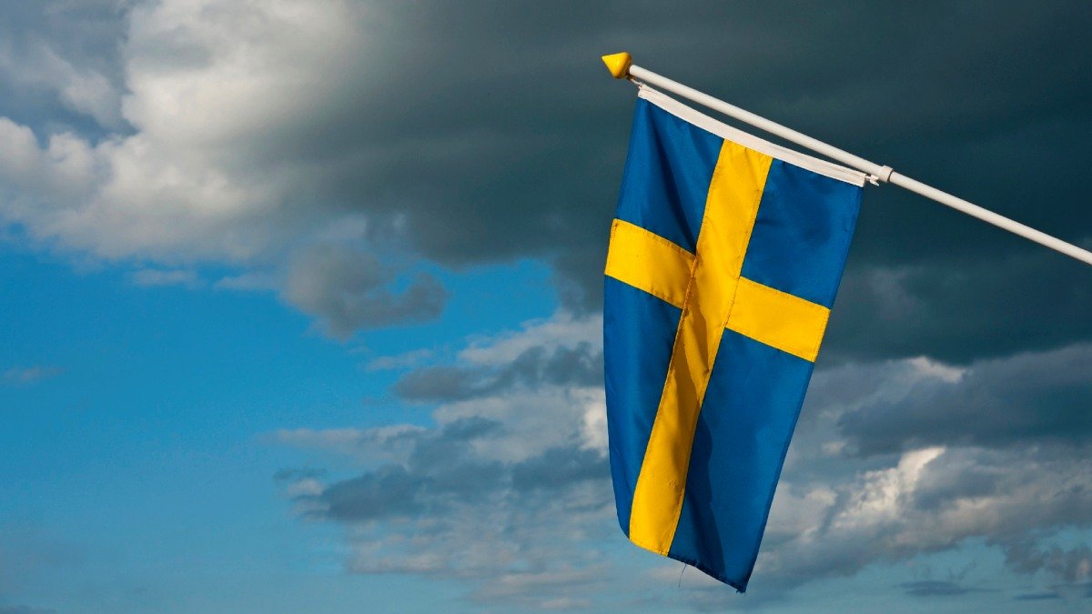 En svensk flagga hänger i luften, bakom finns grå regnmoln.