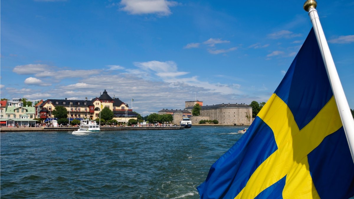 Svensk flagga vajar i förgrunden, från båt, Vaxholm i bakgrunden. Sommar.