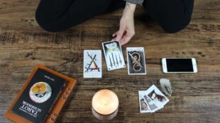 En hand håller i ett tarotkort. Någon sitter på knä på trägolv framför utlagda tarotkort, ett brinnande stearinljus och en bok om tarot.