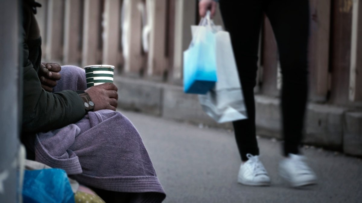 En person som sitter på marken med en burk i handen. En annan person går förbi.