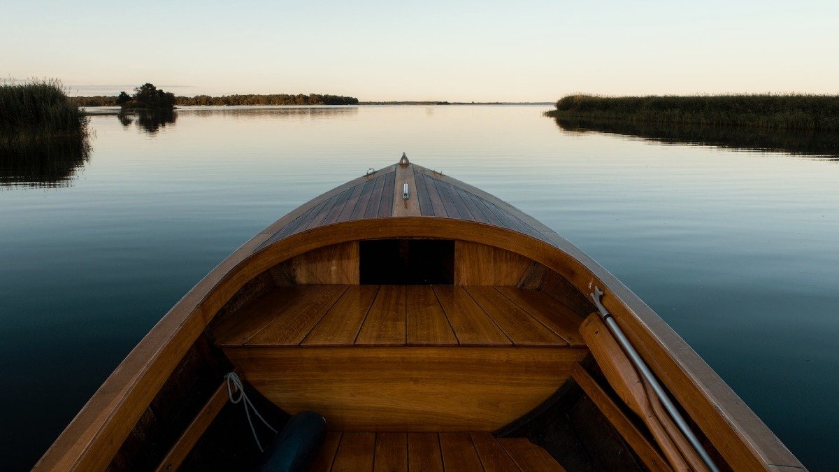 Fören på en brun träbåt, stilla hav och lite land framför.