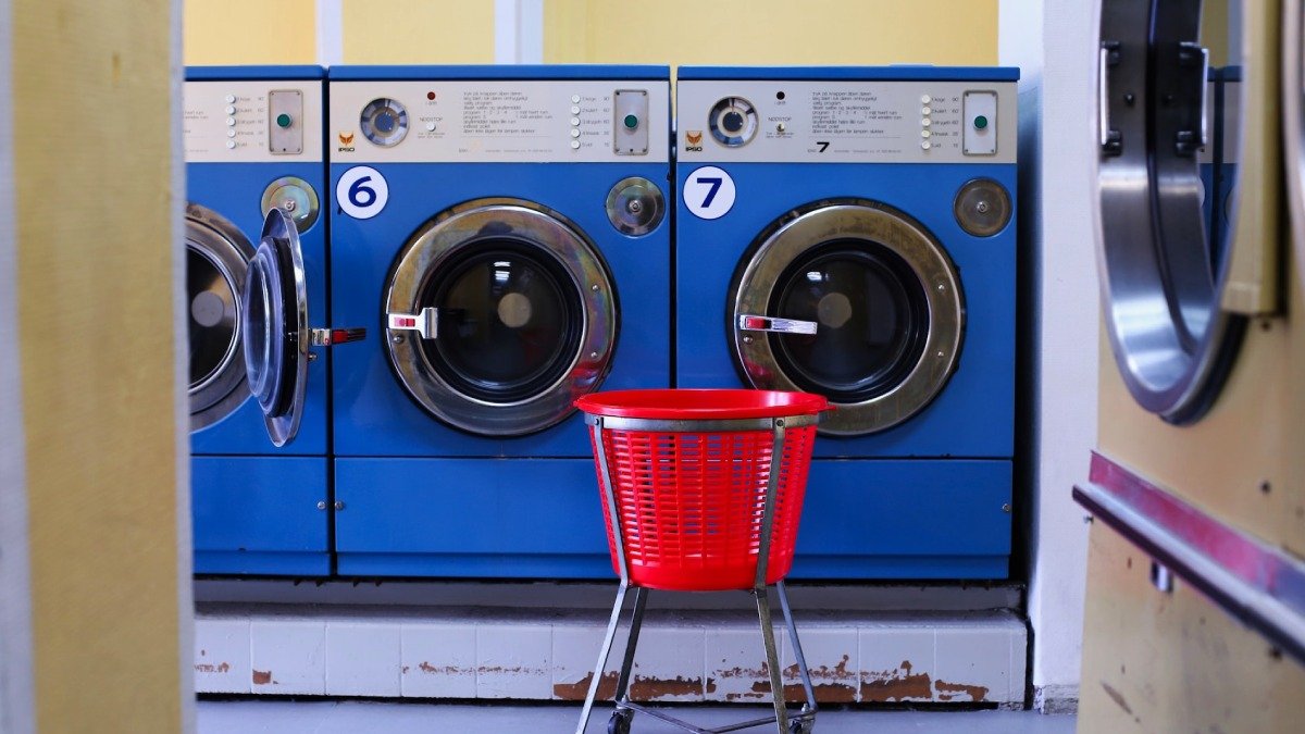Två stora blå tvättmaskiner i tvättstuga, röd plastkorg på hjul framför