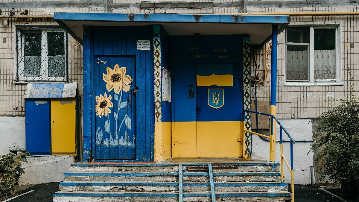 Bild på fasad till ganska nedgången byggnad i Ukraina. Dörrar är målade i blått och gult, med bland annat gula blommor.