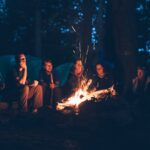 en grupp av vänner sitter runt en eld en mörk natt under camping i skog.