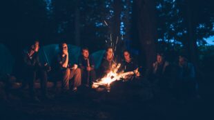 en grupp av vänner sitter runt en eld en mörk natt under camping i skog.