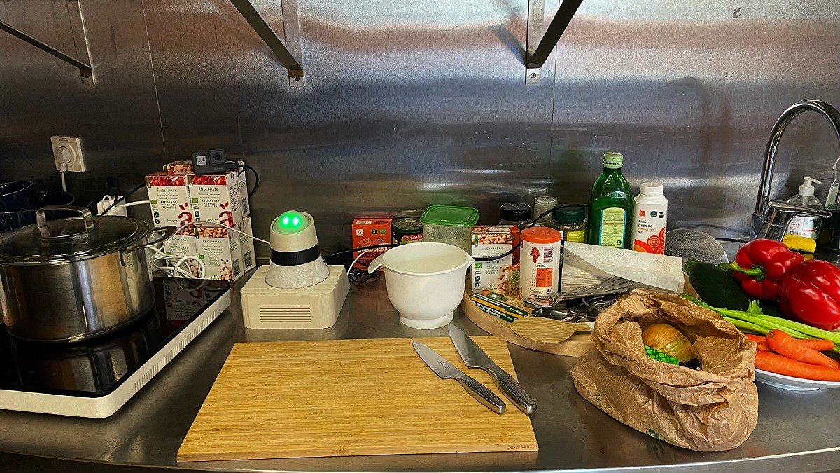 En köksbänk med flaskor och annat. I mitten syns en liten vit robot med gröna ögon bland alla saker.