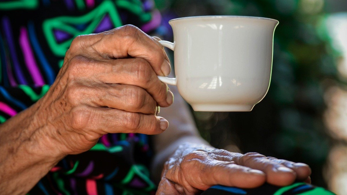 En äldre människas hand håller i en kaffekopp, närbild. Färggranna kläder i bakgrunden.