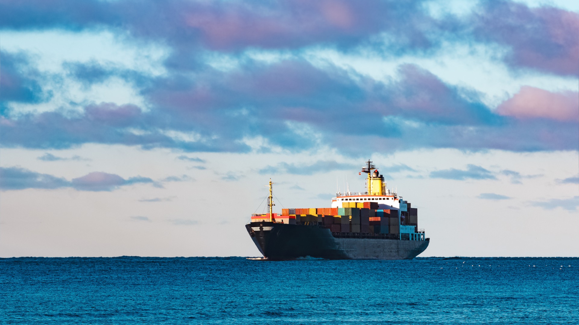 Lastfartyg med containrar syns på avstånd. Blått vatten och blålila moln.