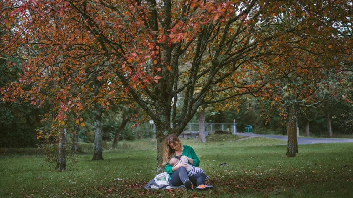 Person med långt hår sitter och ammar bebis i en park med grön gräsmatta och träd med röda löv.