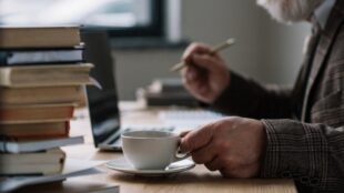 Man med vitt skägg sitter med penna och kaffekopp framför dator. En hög böcker står intill, på bordet.