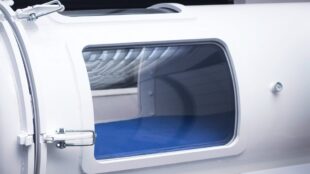 En vit tryckkammare med fönster och blå madrass som syns bakom fönstret.